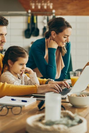 Working father using computer while daughter is sitting on his laptop at home. Mother and daughter are doing homework in the background.